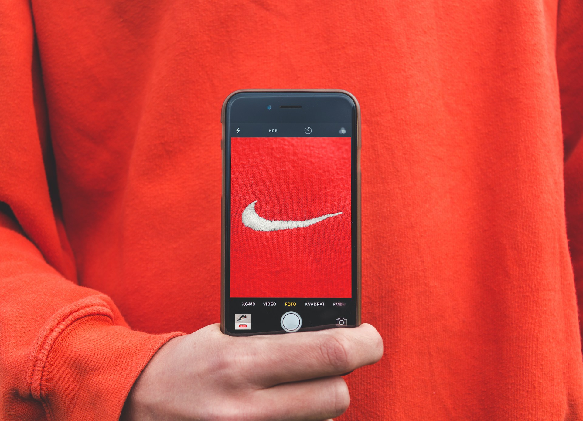 Un telefono fotografa una felpa rossa con il logo Nike indossata. Il logo bianco su sfondo rosso trasmette un'atmosfera sportiva e di stile, evidenziando l'iconicità del marchio Nike.