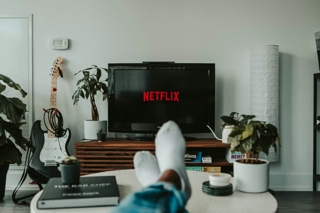 immagine con in mezzo una televisione con il logo netflix nello schermo, dei piedi di una persona che indossa delle calze che guarda la tv. di fianco una chitarra e varie piante