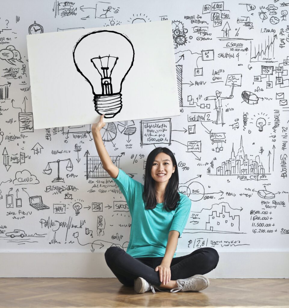 donna che e seduta, mentre tiene in mano un disegno di una lampadina, e lo sfondo dietro alla donna e pieno di disegni di ogni tipo, inerenti al marketing.